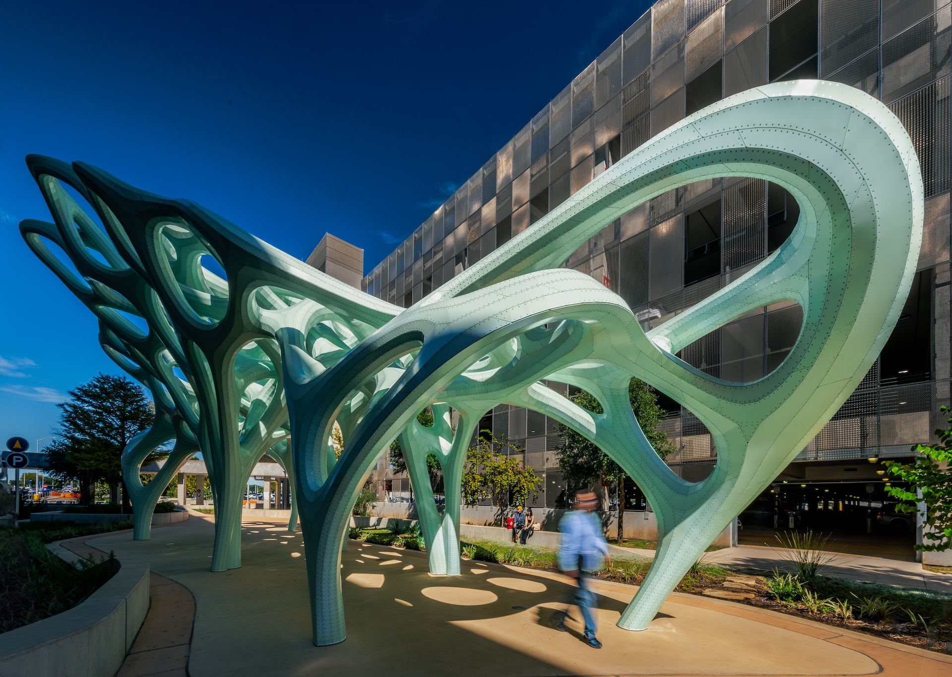 Photograph of large scale light green sculpture with person walking underneath outside the Austin-Bergstrom Airport.