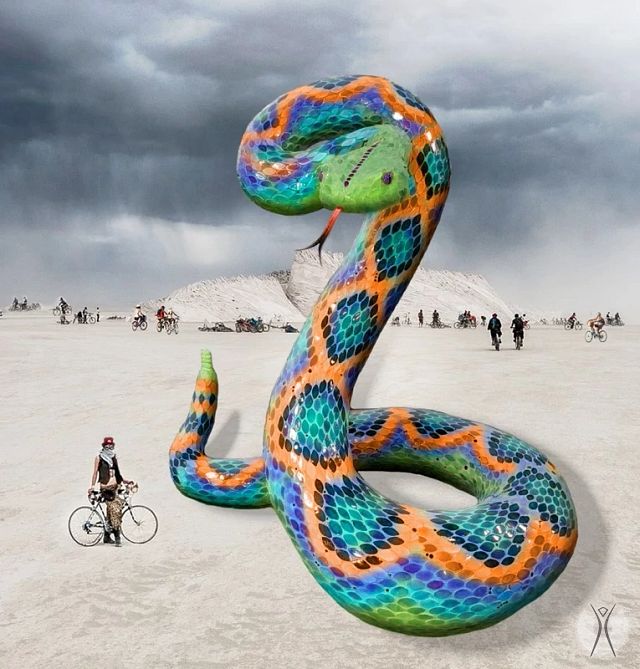 A huge colorful snake sculpture placed outside in the desert with a person on their bike looking at the sculpture.