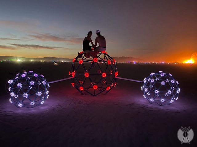 Two people holding hands outside sitting on top of two circular metal-like sculptures watching the sunset.