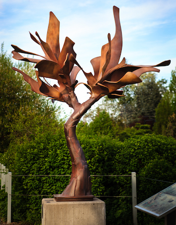 A bronze metal-like tree sculpture standing tall in a park.