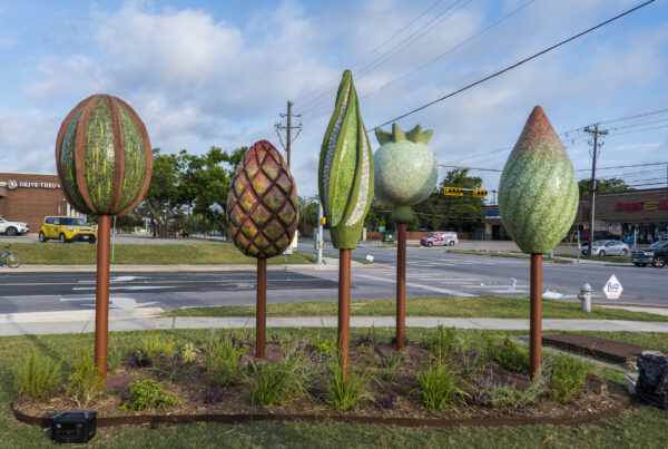 Four large, colorful sculptures resembling fruits or seeds stand in a row on a grassy traffic island at a street intersection. The sculptures are made of mosaic tiles, each with a different shape and color. The sculpture at the far left is round and green with brown stripes. The second sculpture is a brown and orange pinecone shape. The third sculpture is green and looks like a long, pointed seed pod. The fourth sculpture is a green teardrop shape. The sculptures are all attached to rust-colored metal poles. In the background, there are buildings, trees, and traffic. The sky is blue with a few white clouds.