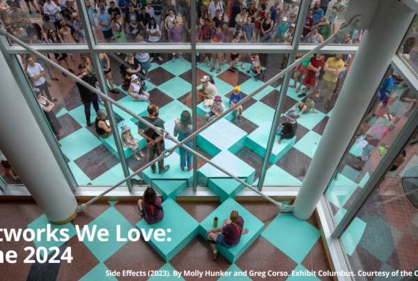 A view from above looking down onto an outdoor art installation. The installation is a series of teal-colored square blocks arranged in a checkerboard pattern. People are sitting on the blocks, interacting with the art piece. There are also many people standing behind a glass window looking down at the installation. The text "Artworks We Love: June 2024" is displayed in white letters on a blue and brown background. Below the text, it reads "Side Effects (2023). By Molly Hunker and Greg Corso. Exhibit Columbus. Courtesy of the Collection."