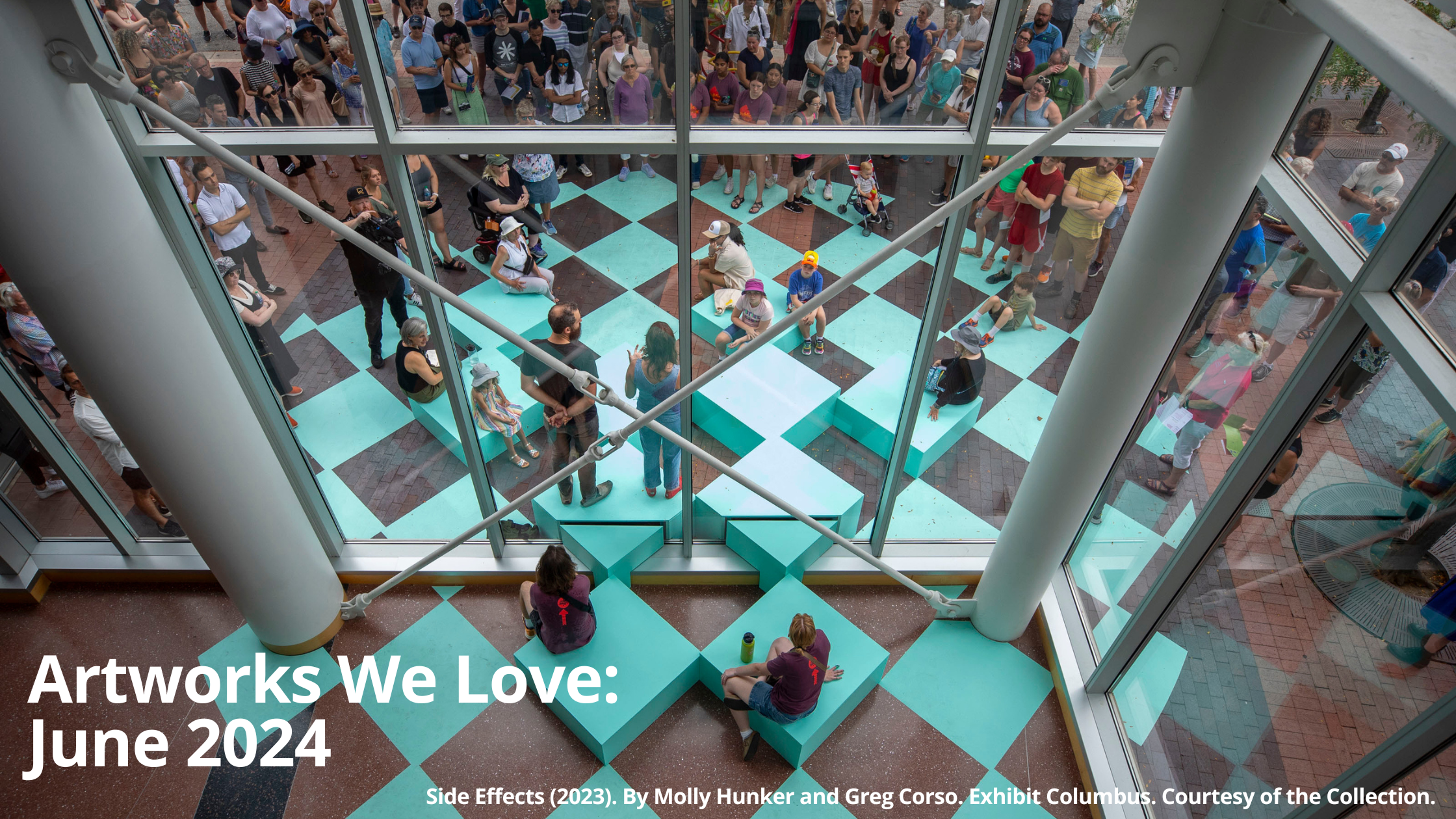 A view from above looking down onto an outdoor art installation. The installation is a series of teal-colored square blocks arranged in a checkerboard pattern. People are sitting on the blocks, interacting with the art piece. There are also many people standing behind a glass window looking down at the installation. The text 