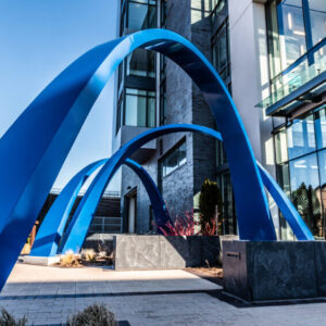 A blue sculpture with three arches, positioned in front of a building