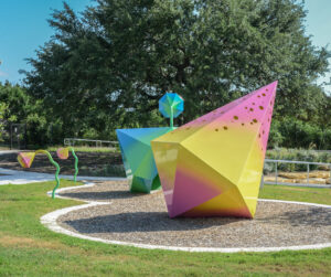 Colorful geometric sculptures in a park setting with trees in the background.