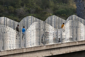 Curved metal panels with vertical slits forming a fence along an elevated pathway.