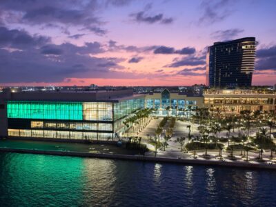 Image at sunset with large glass facade perched near water. Green light emits from the building and palm trees line the interior area around building.