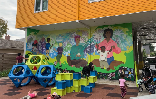 Photograph of building with a colorful painted on it. In front of mural there are rectangular blue and lime green spherical and box-like playground equipment.