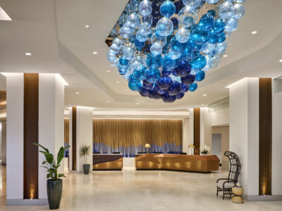 Image of interior space with white walls and wooden panels centered within large white columns. A large sculpture is installed in the center of the ceiling with many circular glass balls in light blue, cerulean blue, and dark blue that hang in an abstract bulbous shape. In the background a panel wall appears to be made of wooden strips with blue paint towards the bottom of the strips appears behind a wooden, geometric front desk.