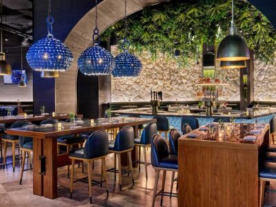 Image of interior of restaurant with blue tile pendant lights hovering over tall rectangualr wooden tables with black bar stools. A green wall composed of plants hangs in the background of the ceiling.