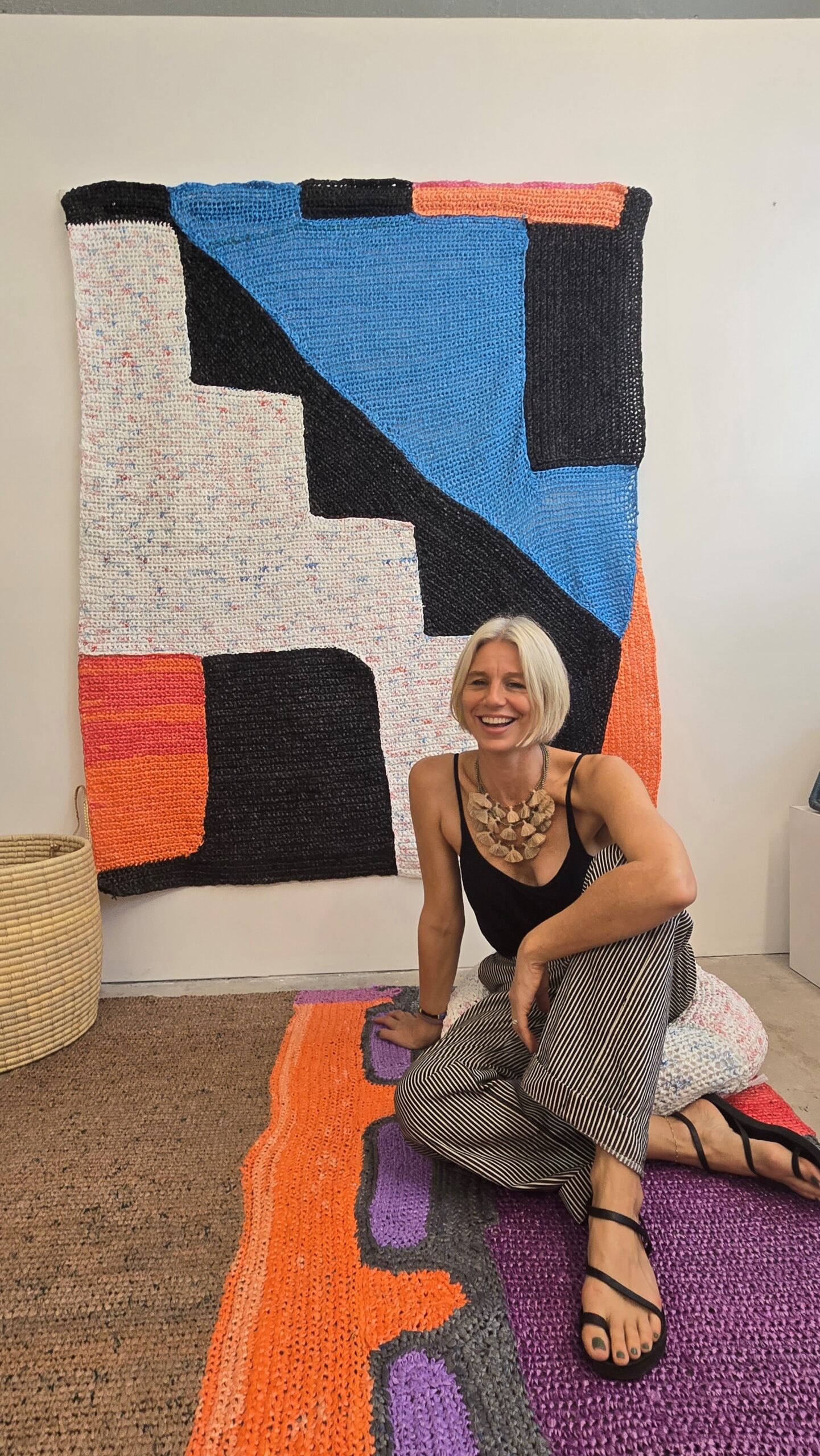 Woman seated in front of large geometric tapestry with blue, black, white and reddish-peach shapes.